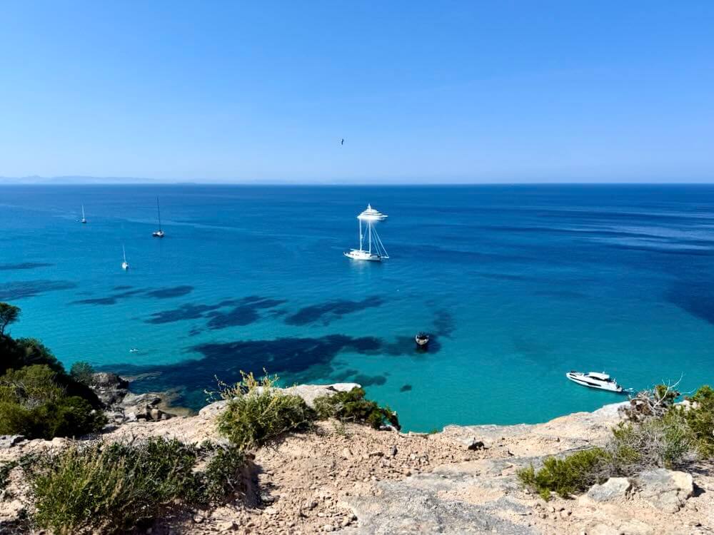 Clear blue waters with several boats anchored near the coastline, framed by lush greenery and a sunny sky on Formentera.