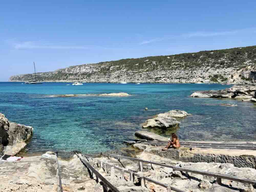 A serene view of turquoise waters, rocky shore, and a person relaxing on the beach in Formentera, Spain.