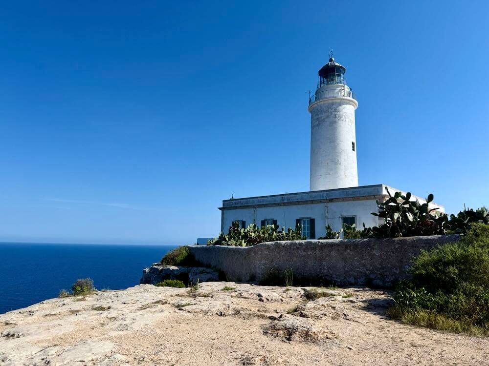 Lighthouse overlooking the Mediterranean Sea on Formentera, surrounded by rocky terrain and cacti under a clear blue sky.