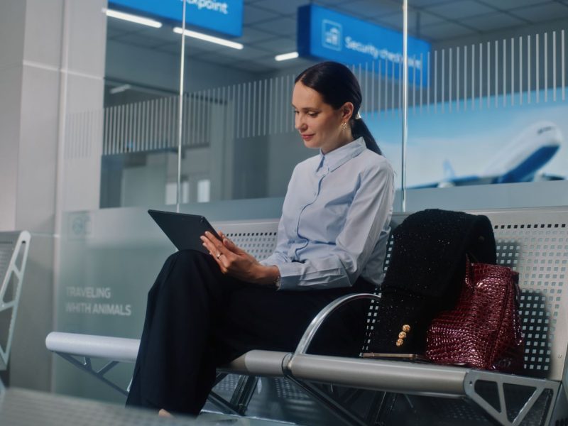Woman using a tablet while waiting at an airport, accompanied by a stylish handbag and a black shawl.