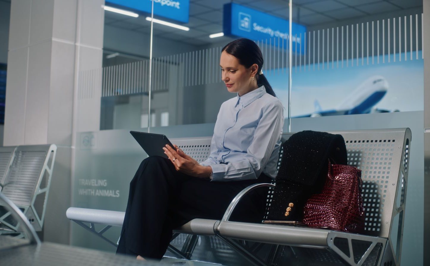 Woman using a tablet while waiting at an airport, accompanied by a stylish handbag and a black shawl.