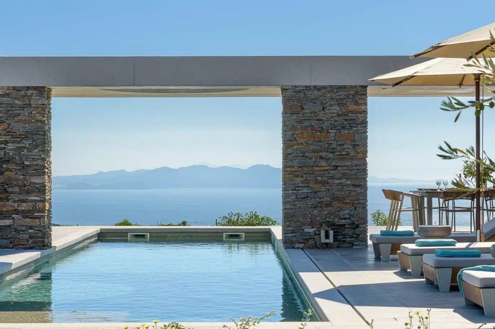Modern outdoor pool with stone columns and a scenic ocean view, featuring lounge chairs and a dining area under umbrellas.