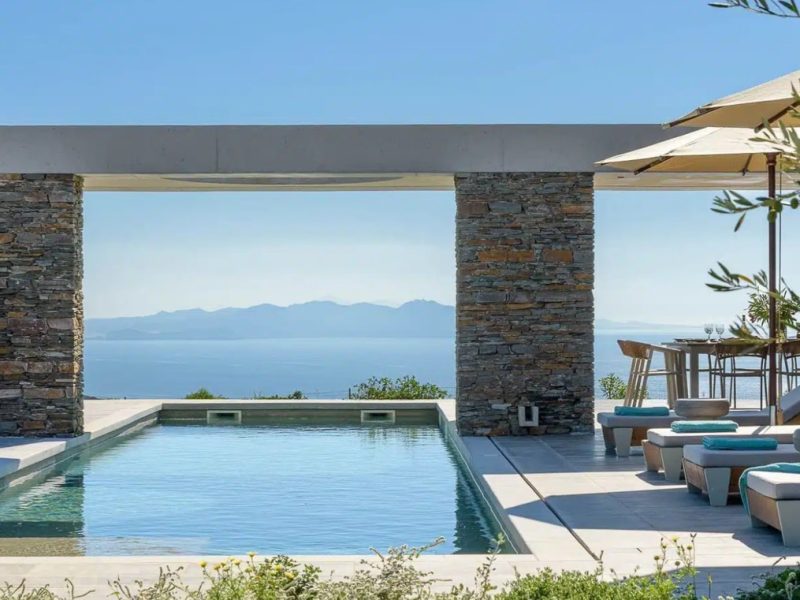 Modern outdoor pool with stone columns and a scenic ocean view, featuring lounge chairs and a dining area under umbrellas.