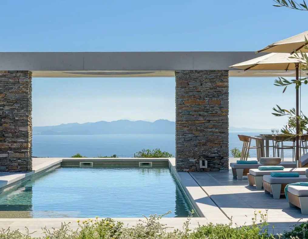 Modern outdoor pool with stone columns and a scenic ocean view, featuring lounge chairs and a dining area under umbrellas.