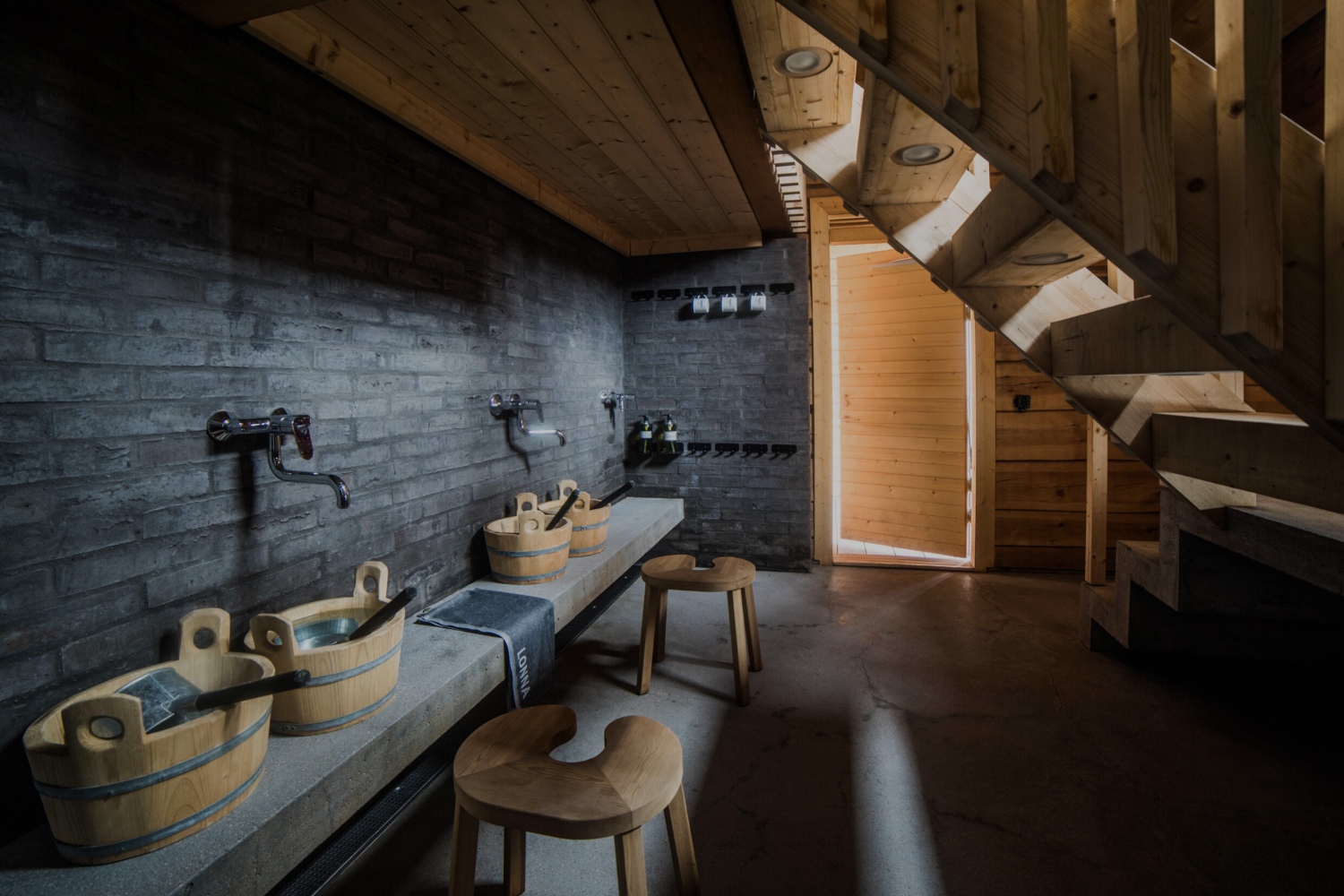 Interior of a sauna featuring wooden tubs, a stone bench, and a staircase leading to a wooden door.