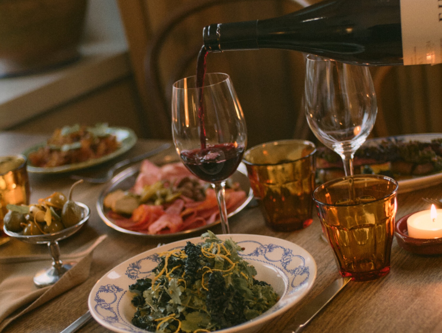 A table set with a variety of dishes, a glass of red wine being poured, and warm candlelight creating a cozy atmosphere.