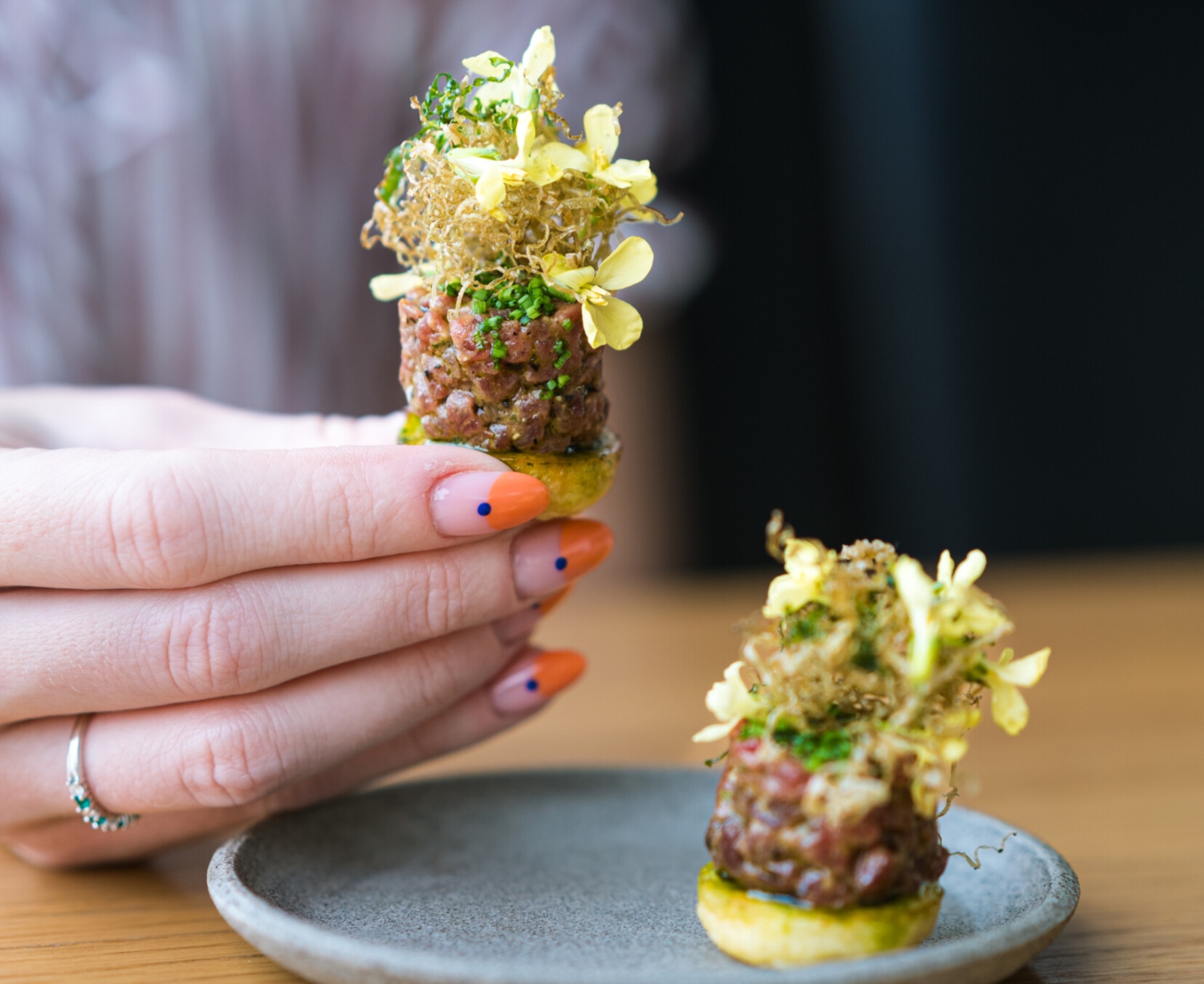 Hand holding a gourmet dish featuring finely chopped meat topped with herbs and edible flowers on a small plate.