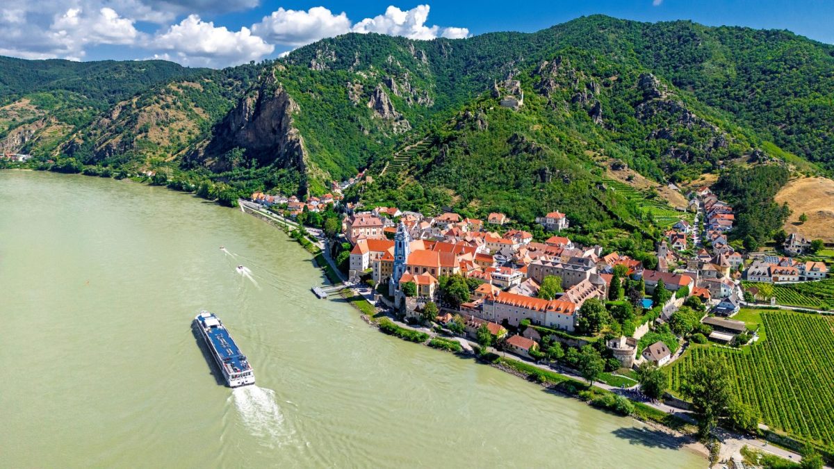 Aerial view of a picturesque riverside town with red-roofed buildings and vineyards, surrounded by lush green hills.