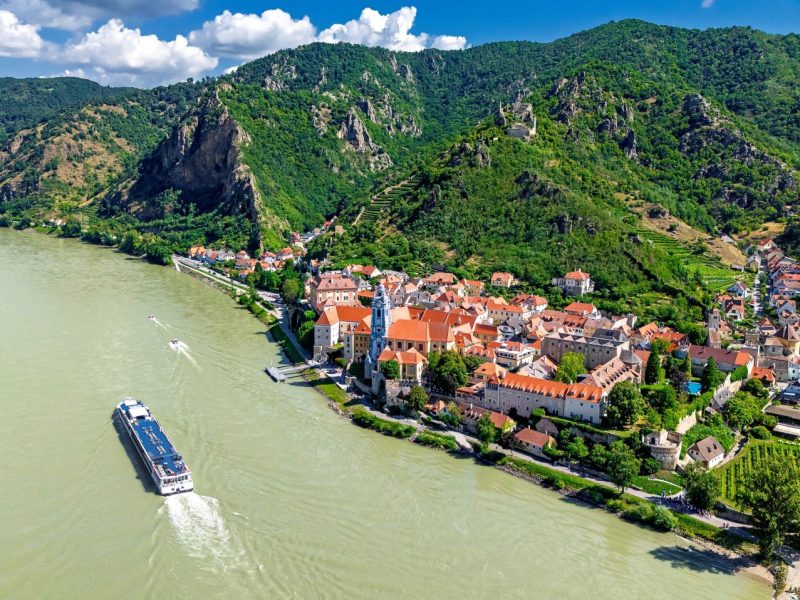 Aerial view of a picturesque riverside town with red-roofed buildings and vineyards, surrounded by lush green hills.