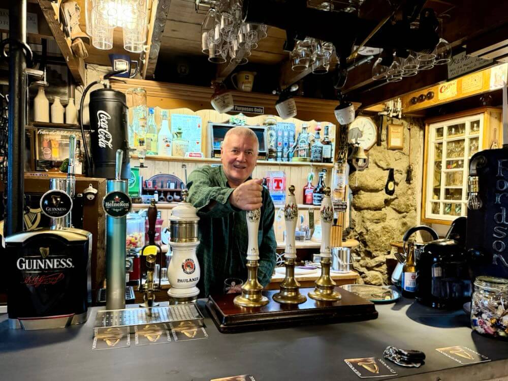 Bartender serving drinks at a well-stocked bar in a luxury Irish guesthouse featuring historic artefacts.
