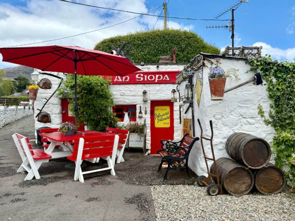 Red cafe tables and chairs outside a white building adorned with historic artefacts and greenery, featuring a striking red umbrella.