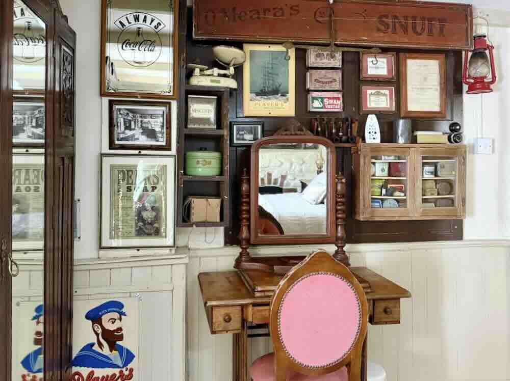 Historic artefacts displayed on a wall, featuring vintage advertisements, a mirror, and a wooden vanity with a pink chair.