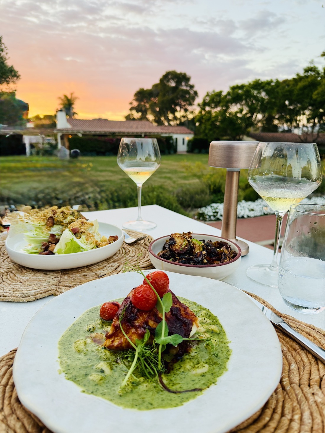 Dinner table set with gourmet dishes and drinks, framed by a vibrant sunset at The Inn at Rancho Santa Fe.