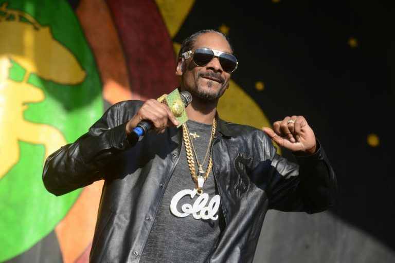 Snoop Dogg performing on stage, wearing sunglasses and a black jacket, with vibrant background elements.