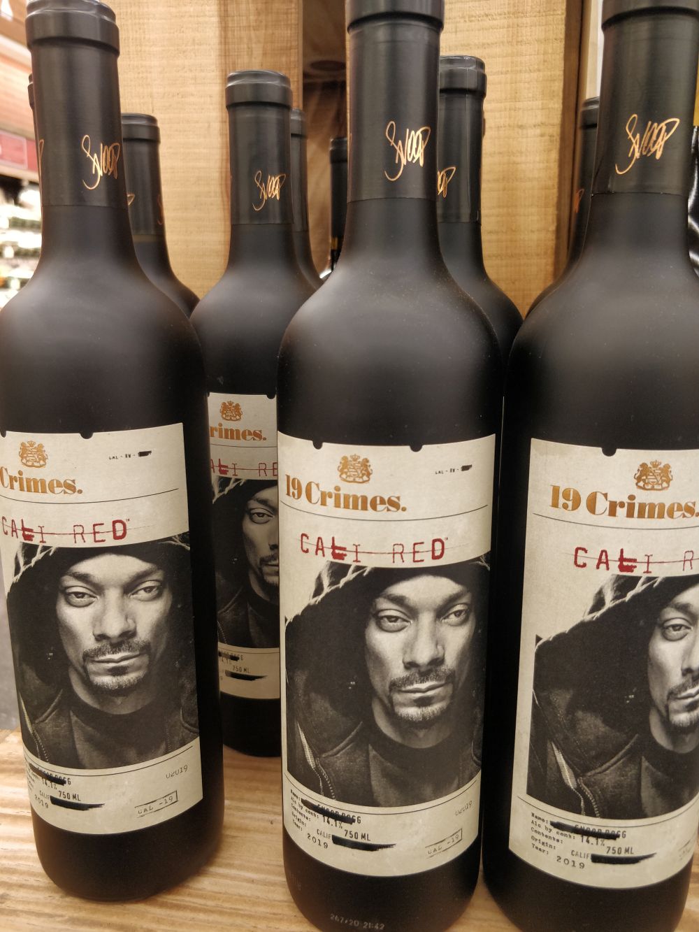 Snoop Dogg's signature on bottles of 19 Crimes Cali Red wine displayed on a wooden shelf.