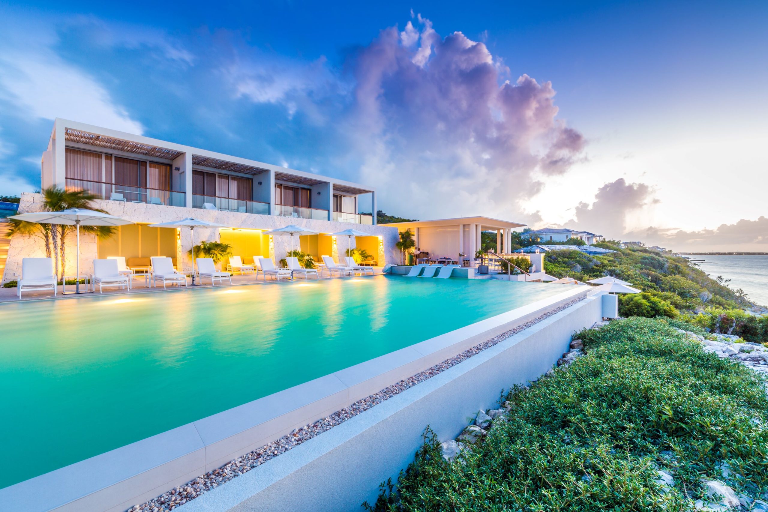 Modern villa with a pool and loungers overlooking the ocean, set against a colorful sunset in Turks and Caicos.