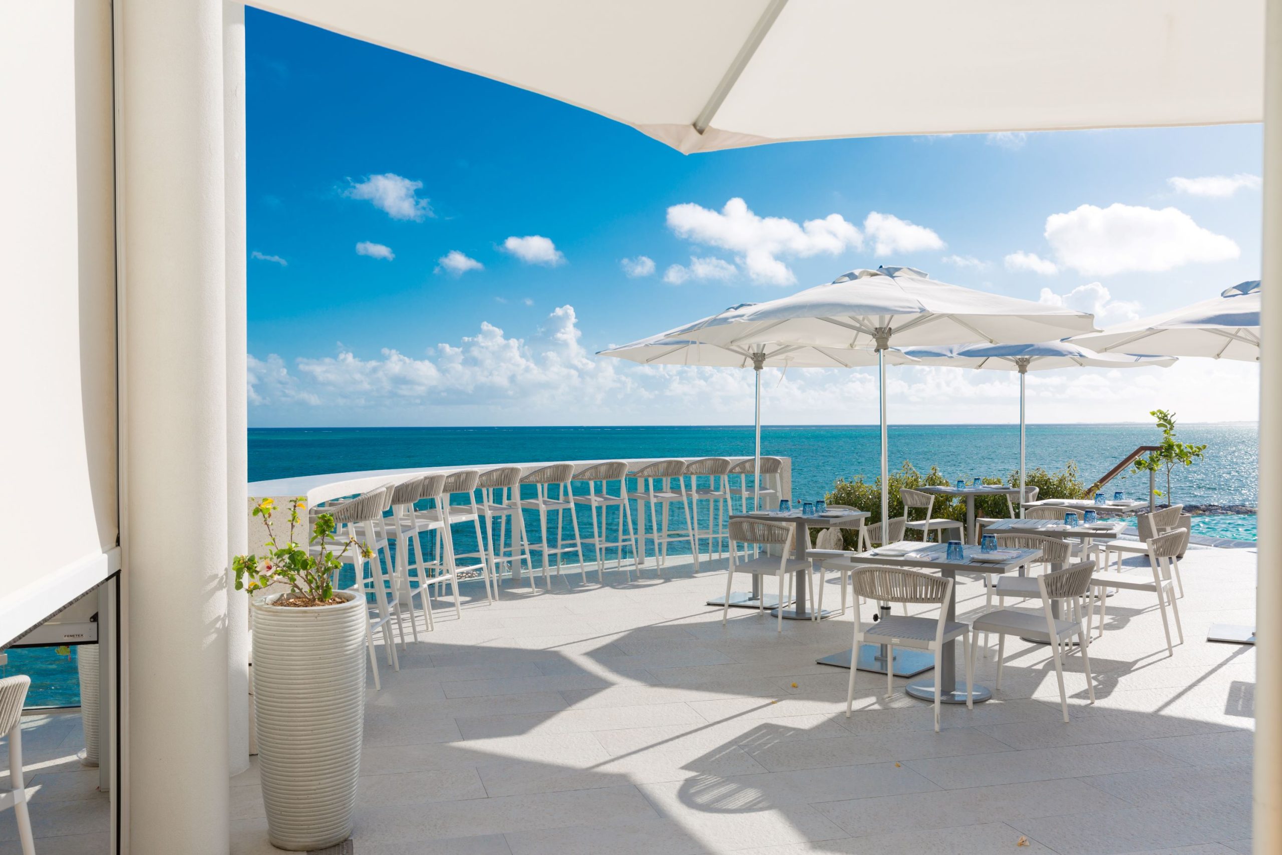 Outdoor dining area with white chairs and umbrellas overlooking the turquoise ocean under a clear blue sky.