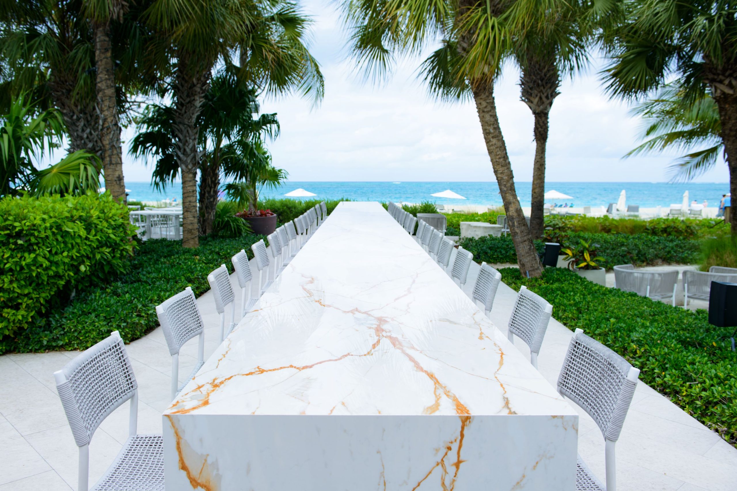 A long marble table with white chairs overlooks the beach, framed by palm trees and greenery in Turks and Caicos.