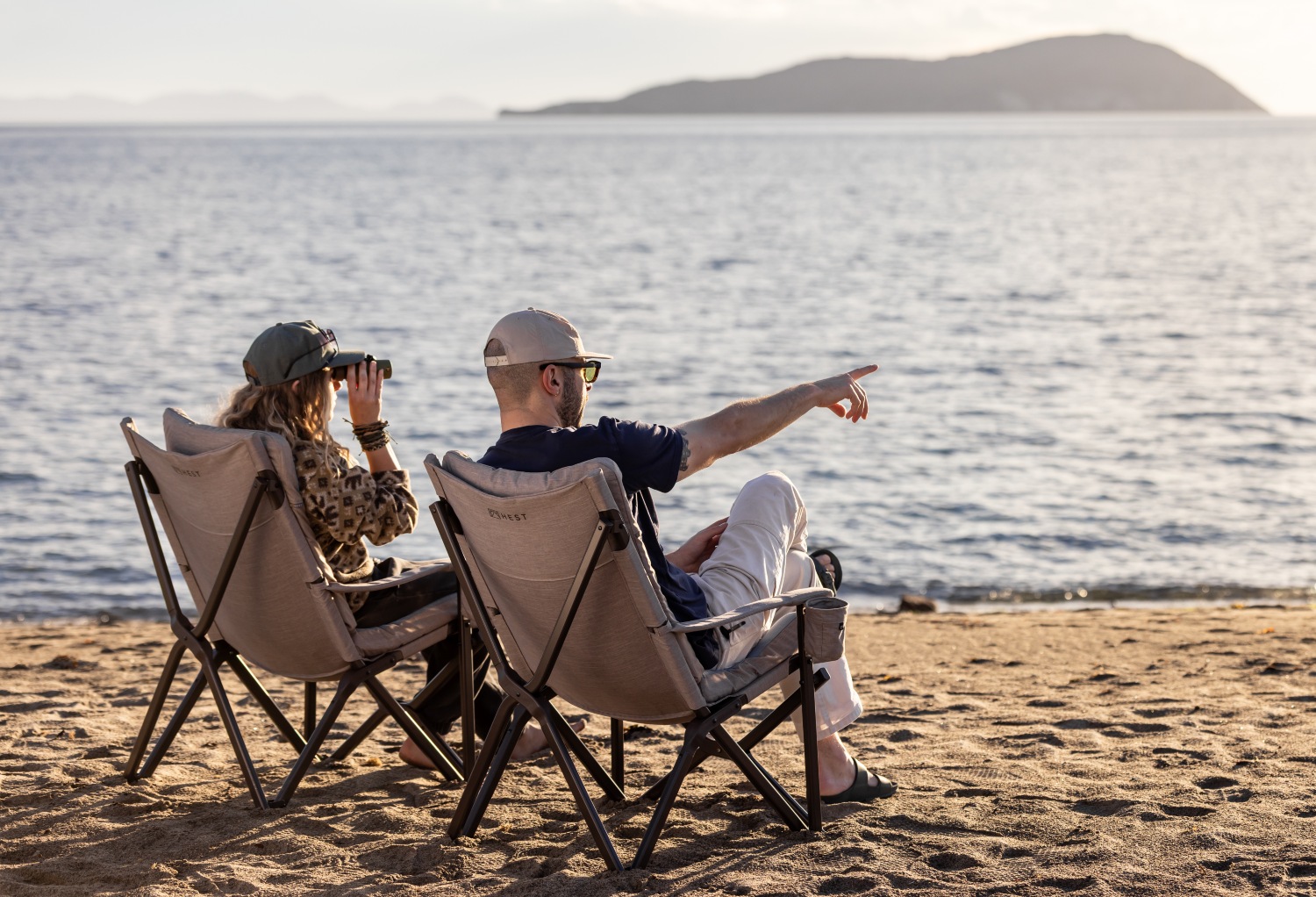A couple relaxes in beach chairs, enjoying the view of the water and pointing towards the horizon.