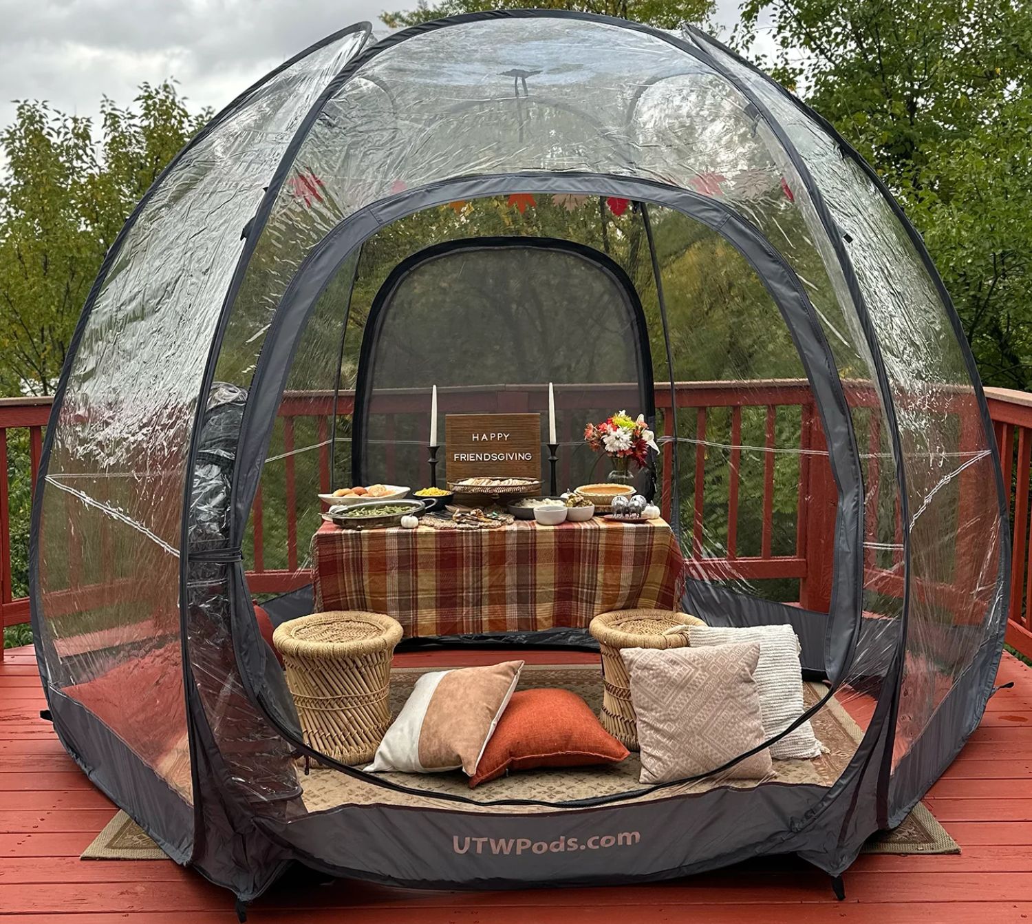 A cozy outdoor setup featuring a clear enclosure with a table of festive food, blankets, and cushions for Friendsgiving.