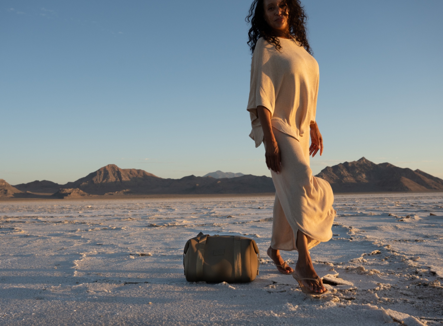 A woman in a beige outfit stands on a salt flat with mountains in the background, next to a stylish travel bag.