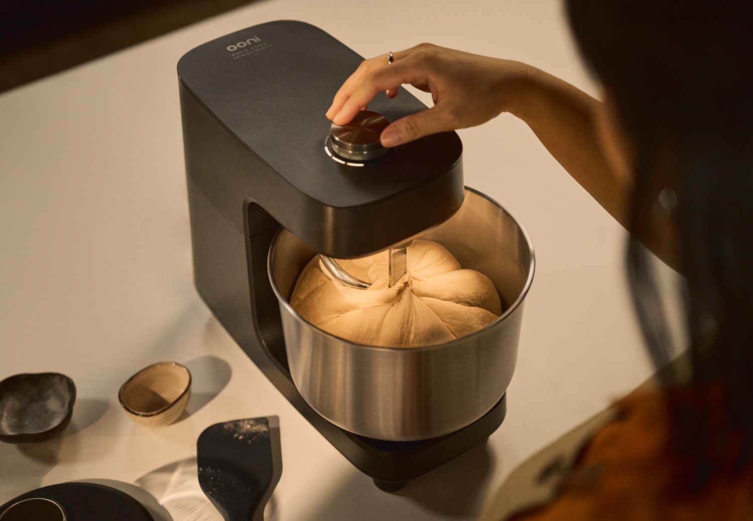 A person using an Ooni mixer with dough inside the stainless steel bowl on a countertop.