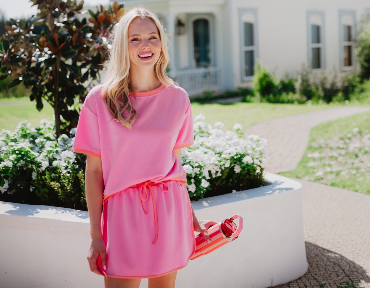 A woman in a bright pink dress smiles while holding a red drink container, surrounded by flowers in a sunny outdoor setting.
