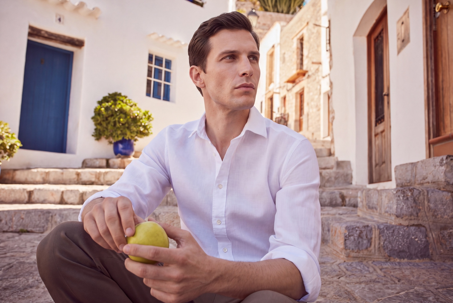 Man sitting on steps holding an apple, wearing a white shirt in a picturesque outdoor setting with white buildings.