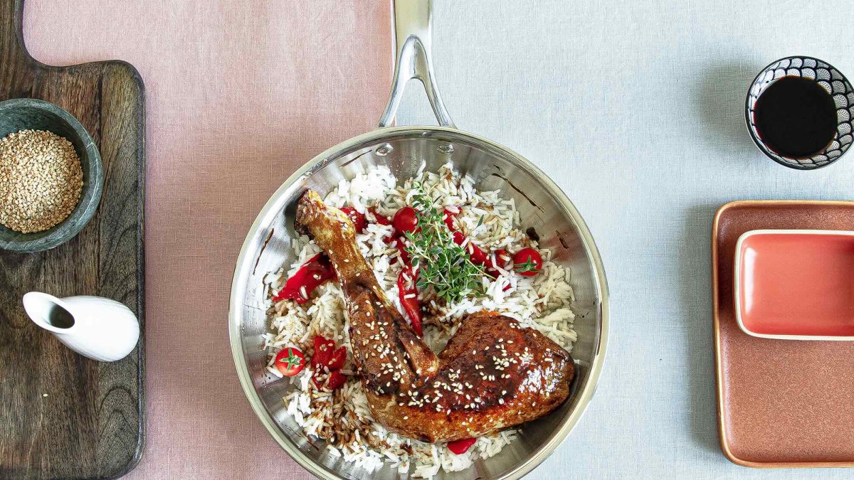A stainless steel frying pan filled with rice, grilled chicken, and garnished with herbs and chili on a colorful table setting.