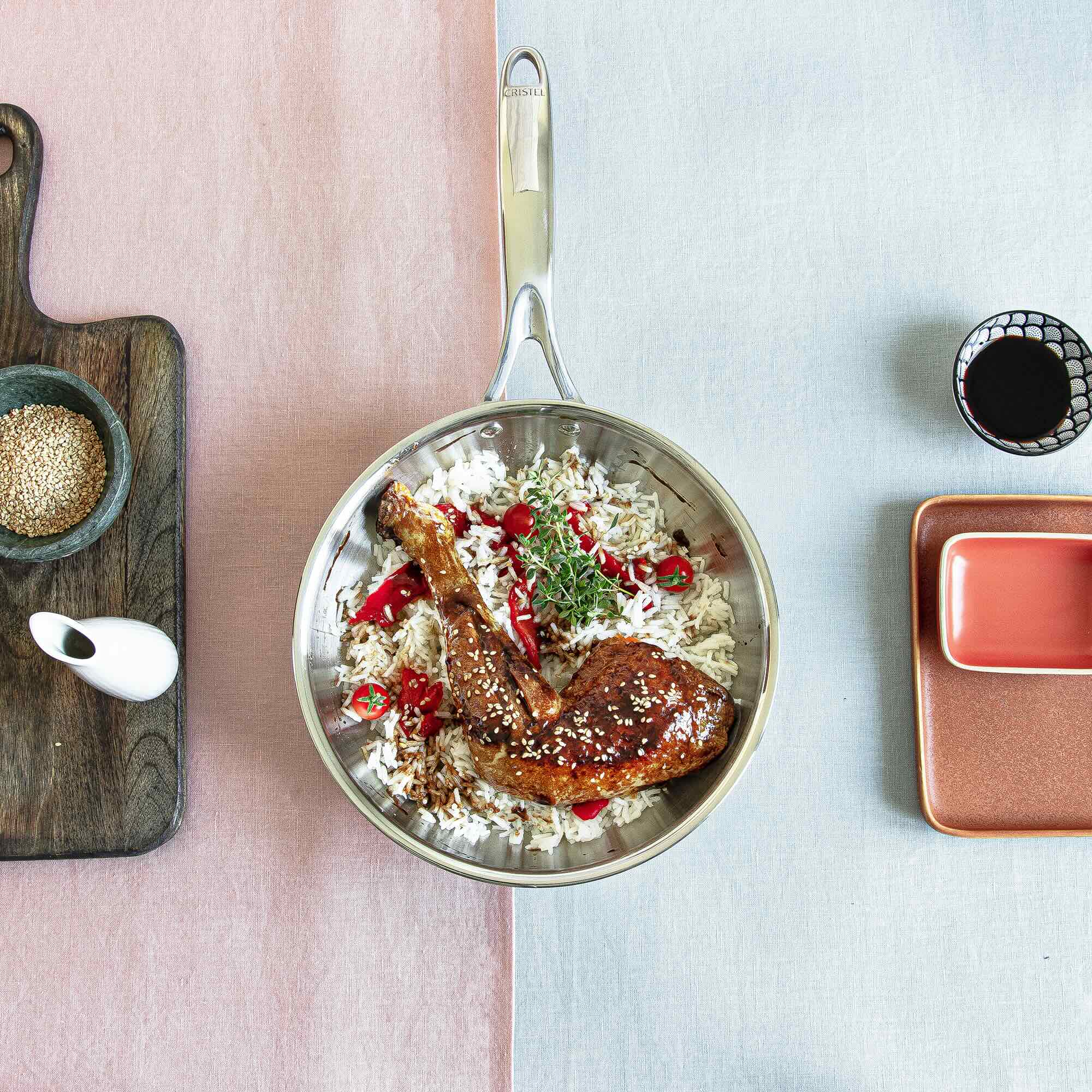 A stainless steel frying pan filled with rice, grilled chicken, and garnished with herbs and chili on a colorful table setting.