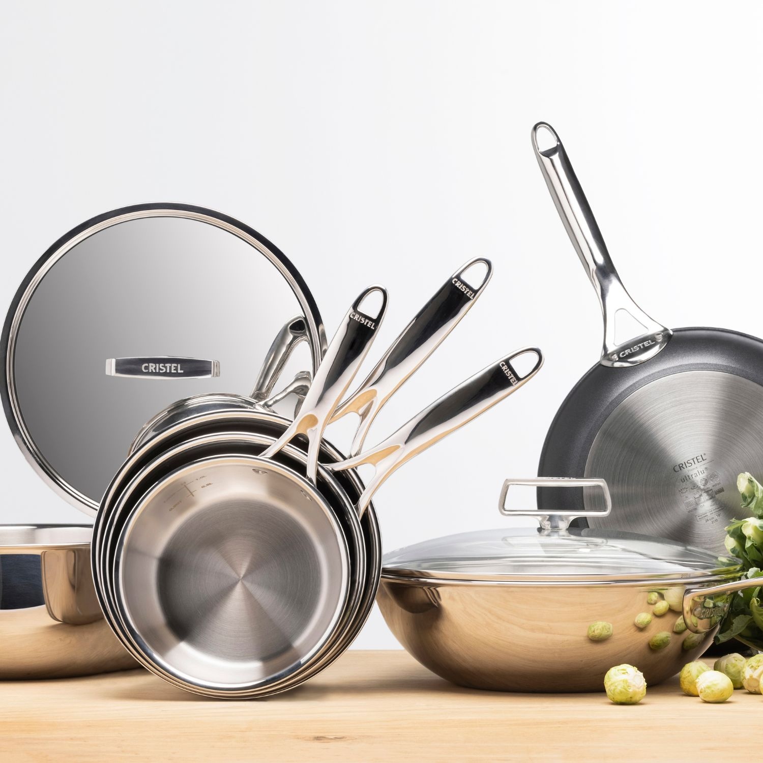 Cristel cookware set featuring stainless steel frying pans, a lid, and a pot, showcased on a wooden surface with Brussels sprouts.