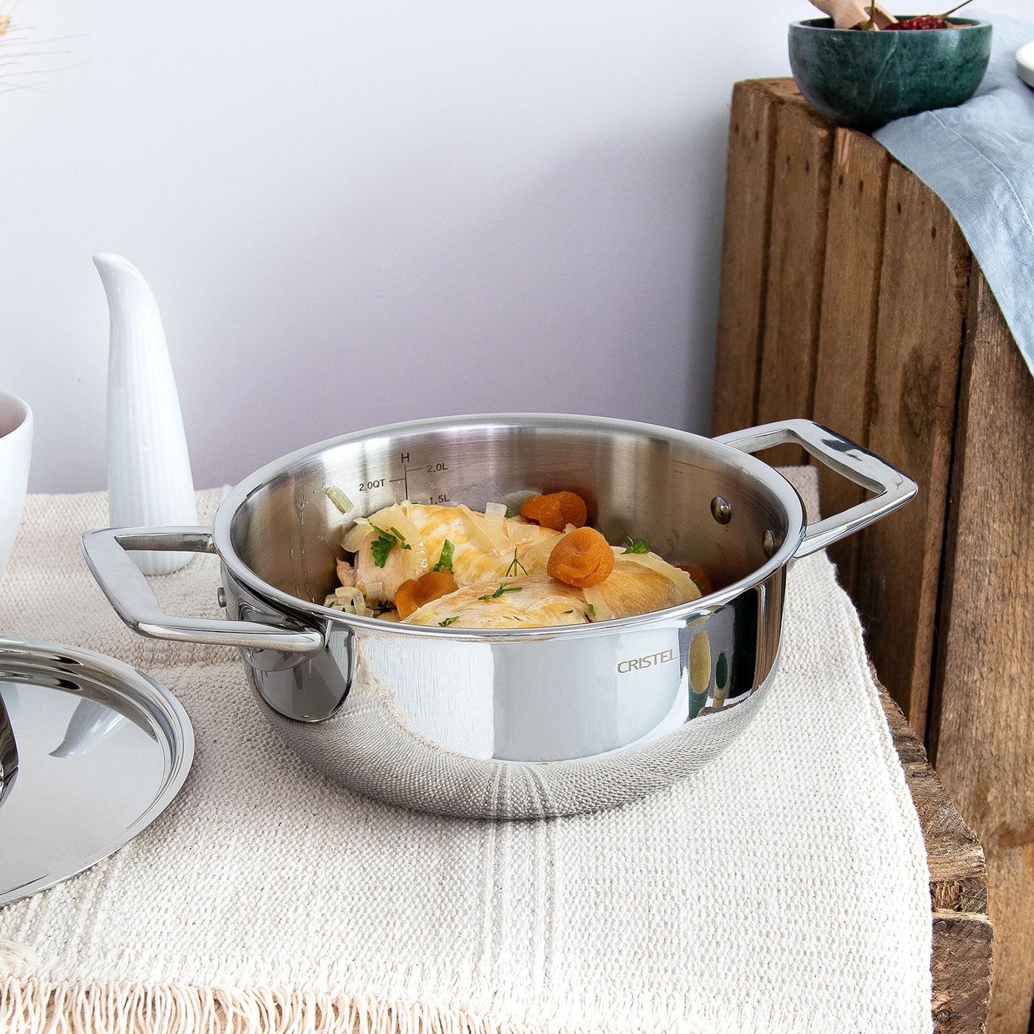 A stainless steel Cristel stewpan with food ingredients, placed on a textured surface, showcasing its reflective finish.