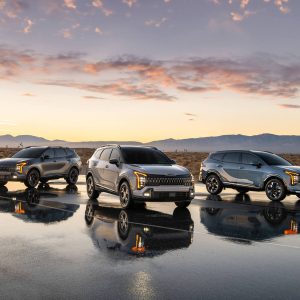 Three Kia SUVs are parked on a reflective surface at sunset, showcasing their sleek designs against a desert backdrop.