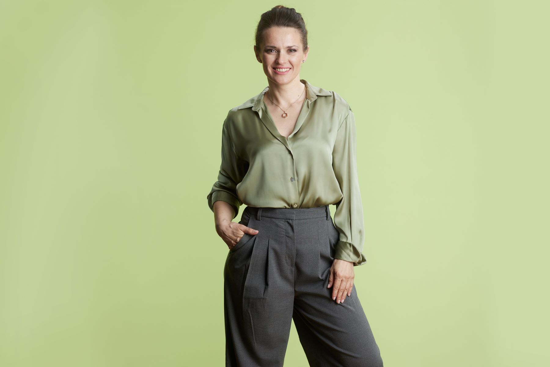 Woman smiling confidently in a green blouse and tailored gray slacks against a light green background.