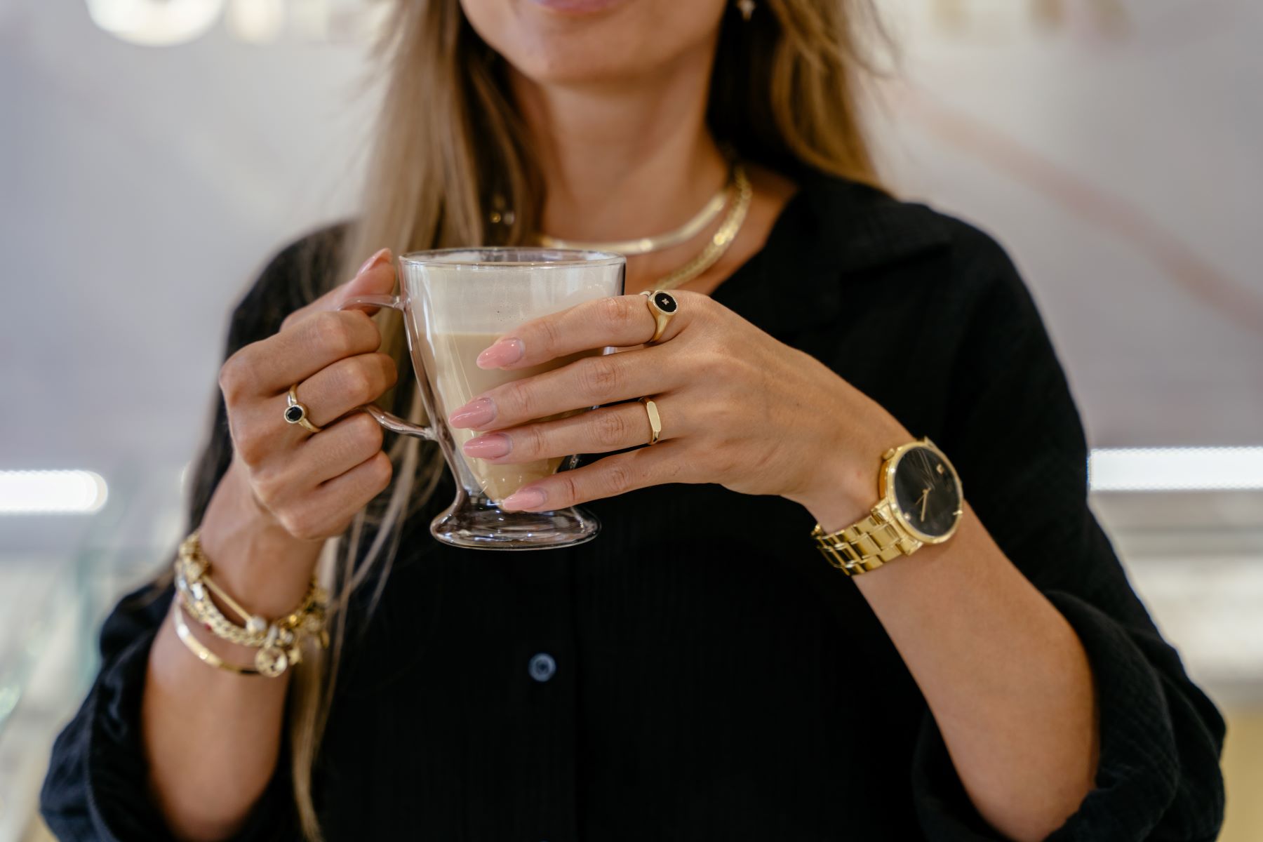 Woman holding a glass of latte, showcasing several rings and bracelets, while wearing a stylish black shirt and gold watch.