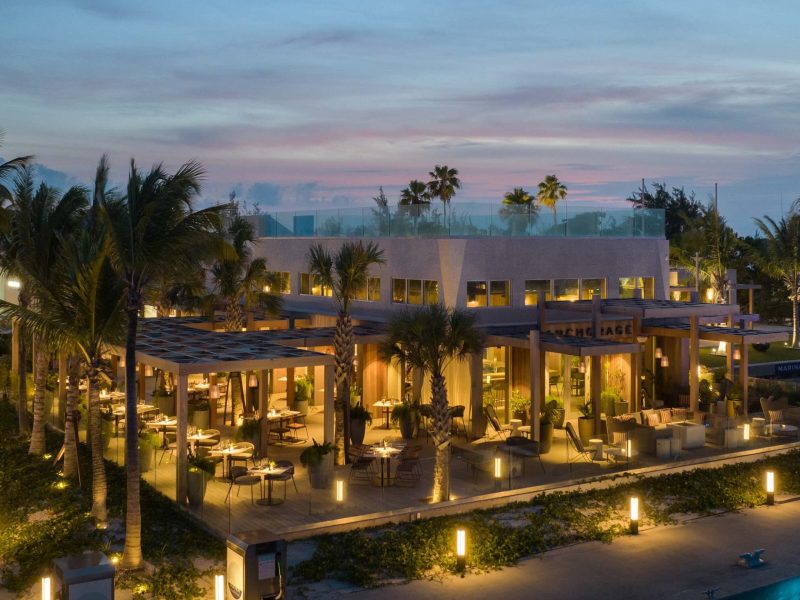 Modern dockside restaurant with outdoor seating, illuminated at dusk, surrounded by palm trees in Turks and Caicos.
