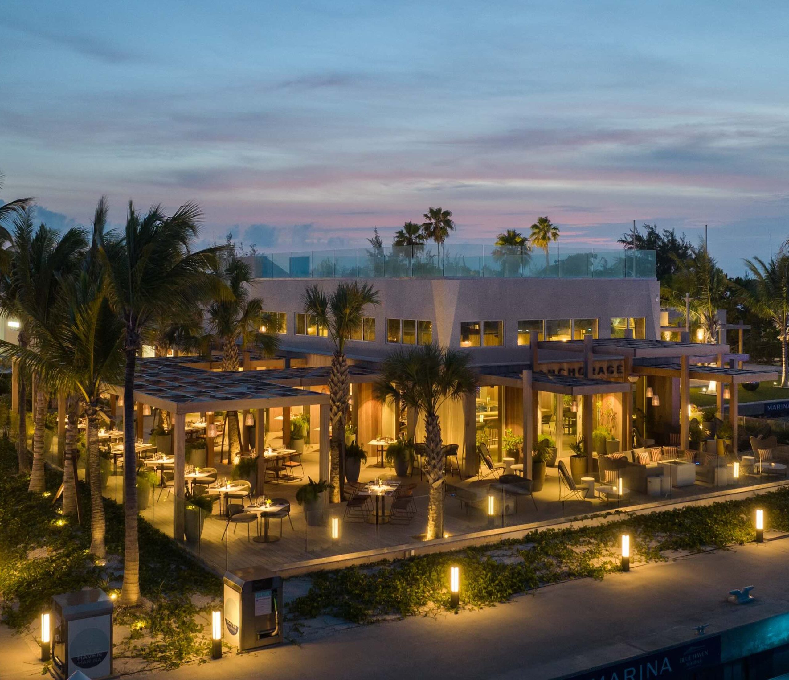 Modern dockside restaurant with outdoor seating, illuminated at dusk, surrounded by palm trees in Turks and Caicos.