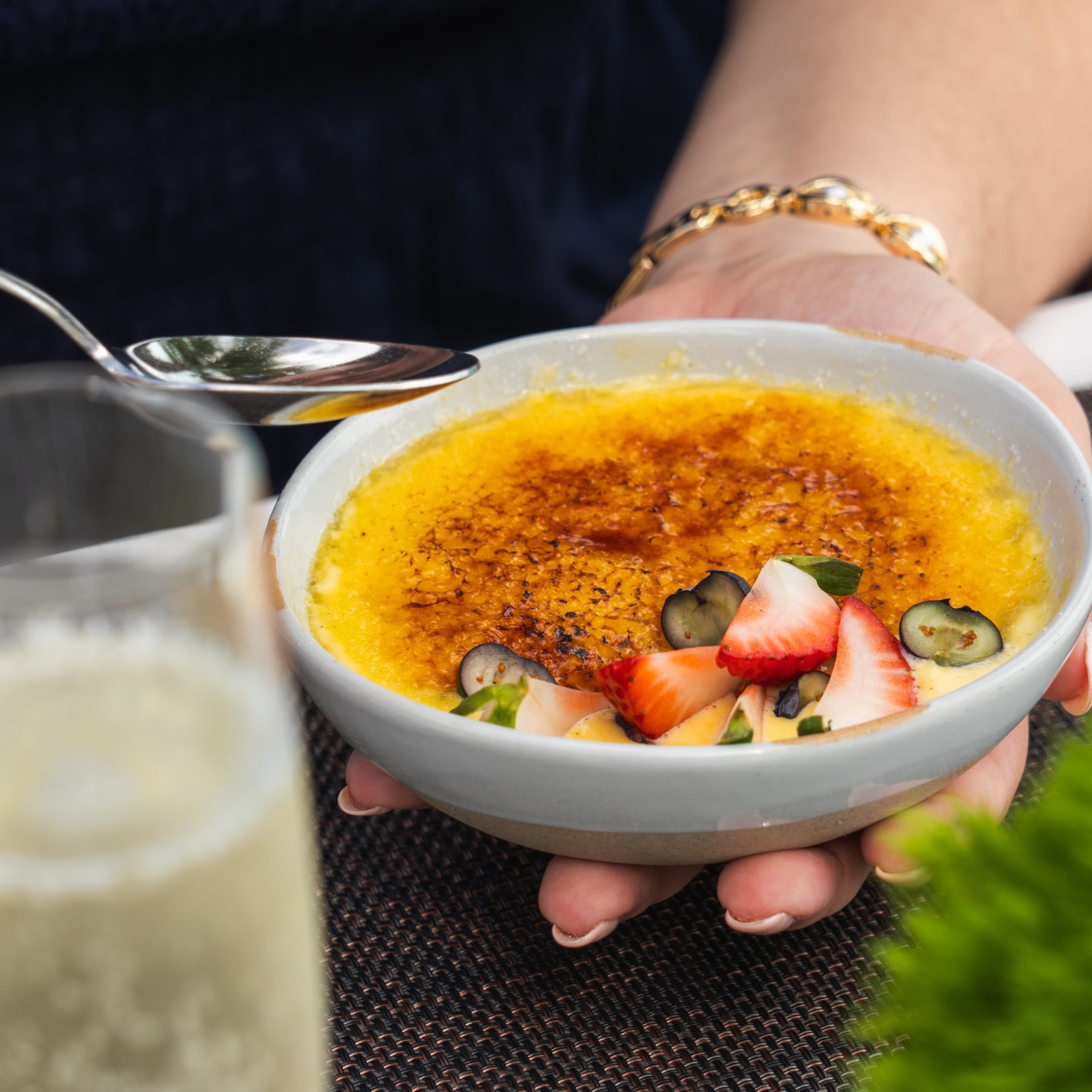 A hand holding a bowl of crème brûlée topped with strawberries and blueberries, with a glass of sparkling drink nearby.