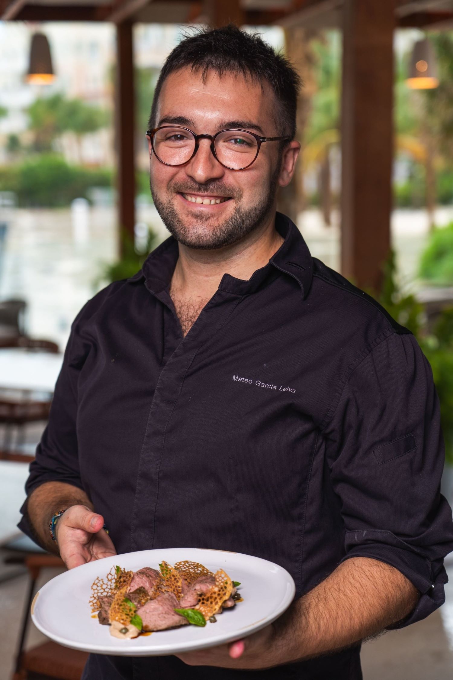 Smiling chef holds a beautifully plated dish featuring meat and decorative elements in a relaxed dining setting.