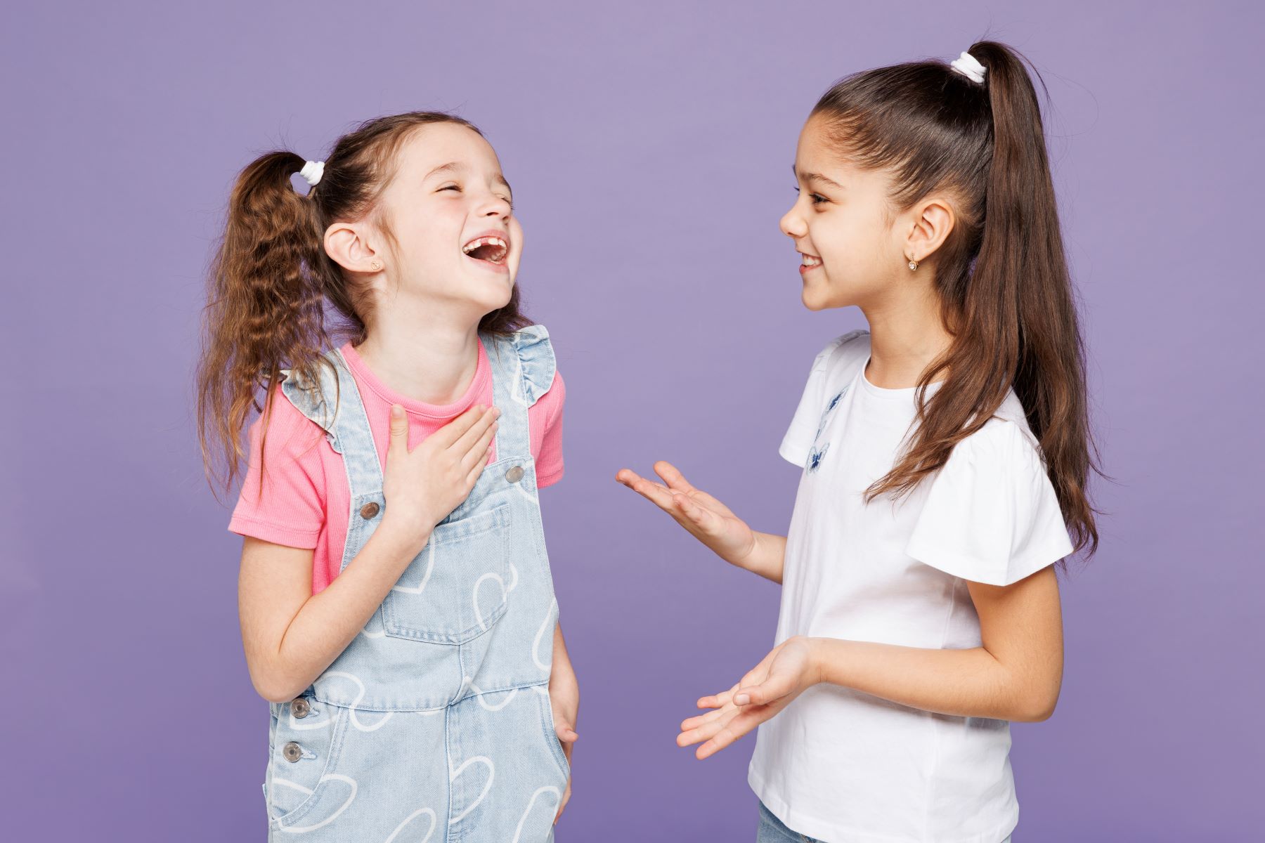 Two girls laughing and chatting in stylish outfits against a purple background, showcasing joy and friendship.