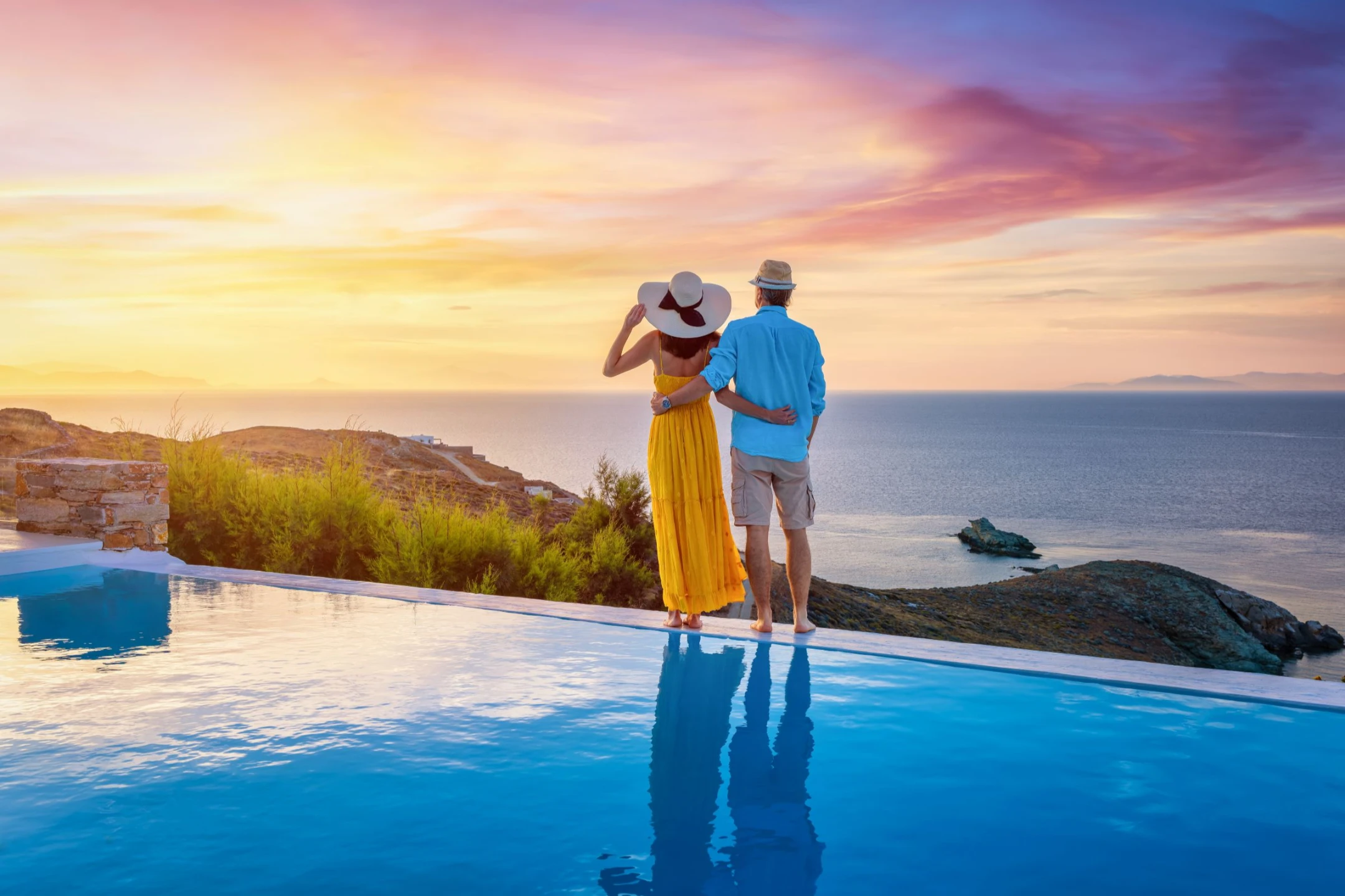 Couple standing by an infinity pool overlooking the sea at sunset, with vibrant colors in the sky.