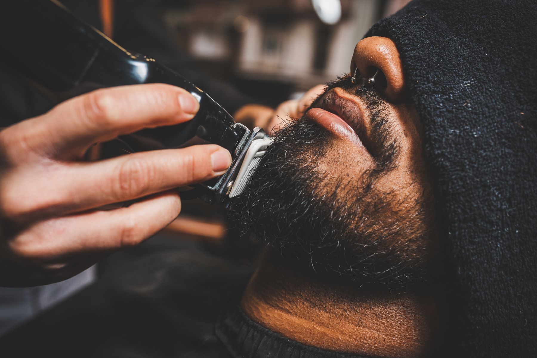 A barber carefully trims a man's beard with clippers, ensuring a clean and precise look in a barbershop setting.