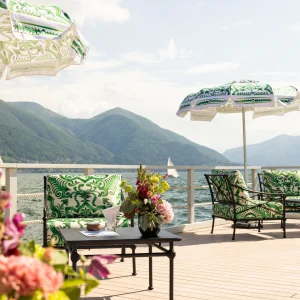 Stylish outdoor seating area with patterned umbrellas and colorful flowers, overlooking a serene lake and mountains.