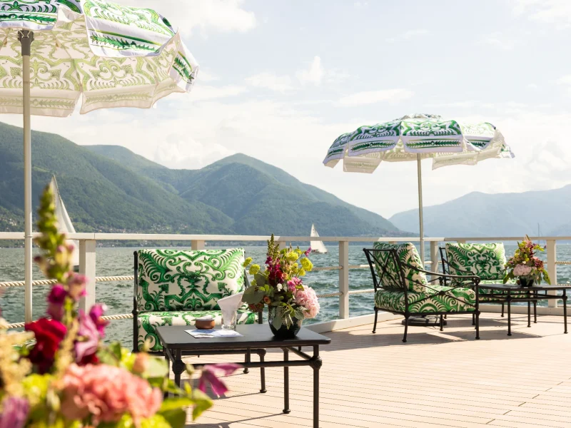 Stylish outdoor seating area with patterned umbrellas and colorful flowers, overlooking a serene lake and mountains.