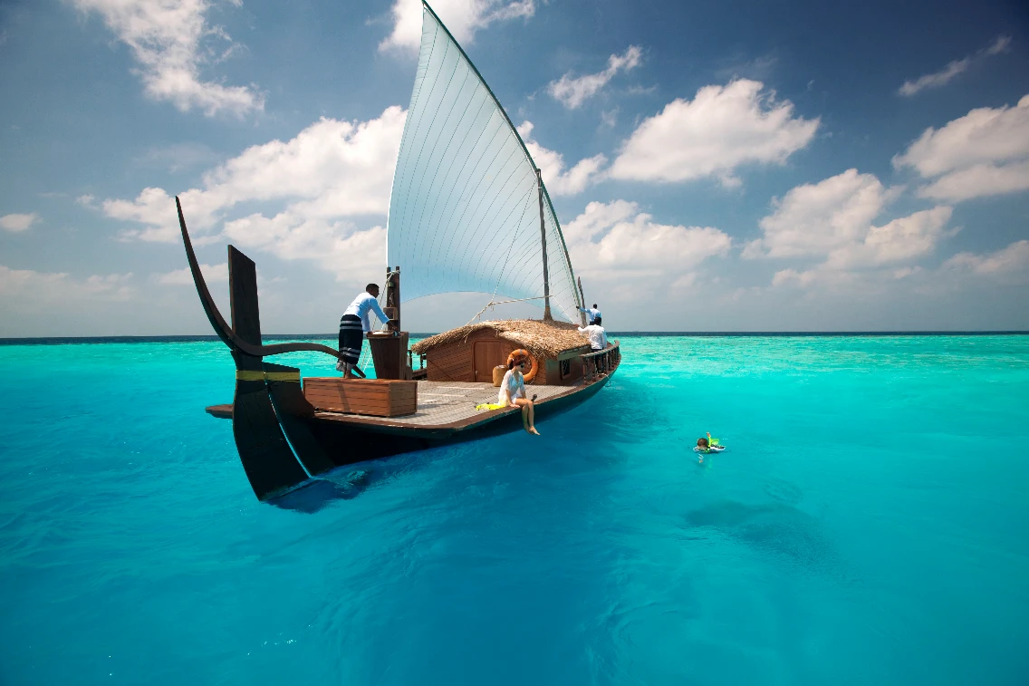 A traditional boat with a sail floats in turquoise waters, while a person relaxes on board and another swims nearby.