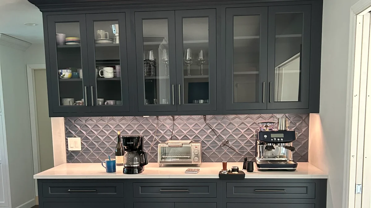 Modern kitchen cabinetry with glass doors, coffee maker, espresso machine, and neutral tile backsplash.