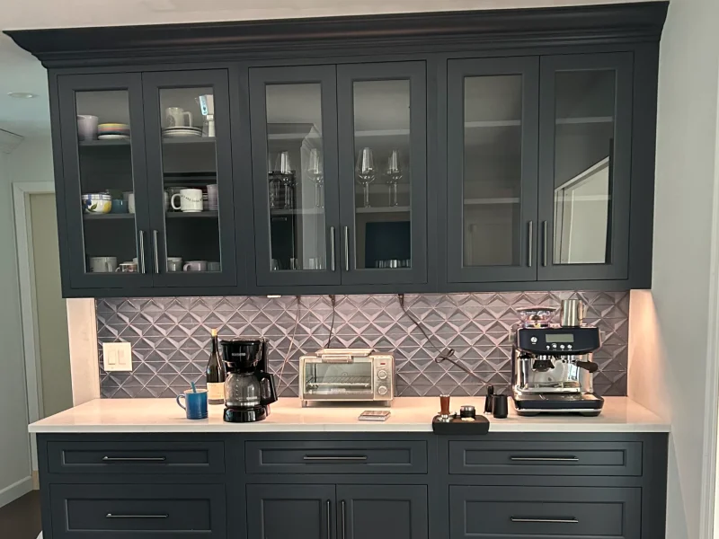 Modern kitchen cabinetry with glass doors, coffee maker, espresso machine, and neutral tile backsplash.