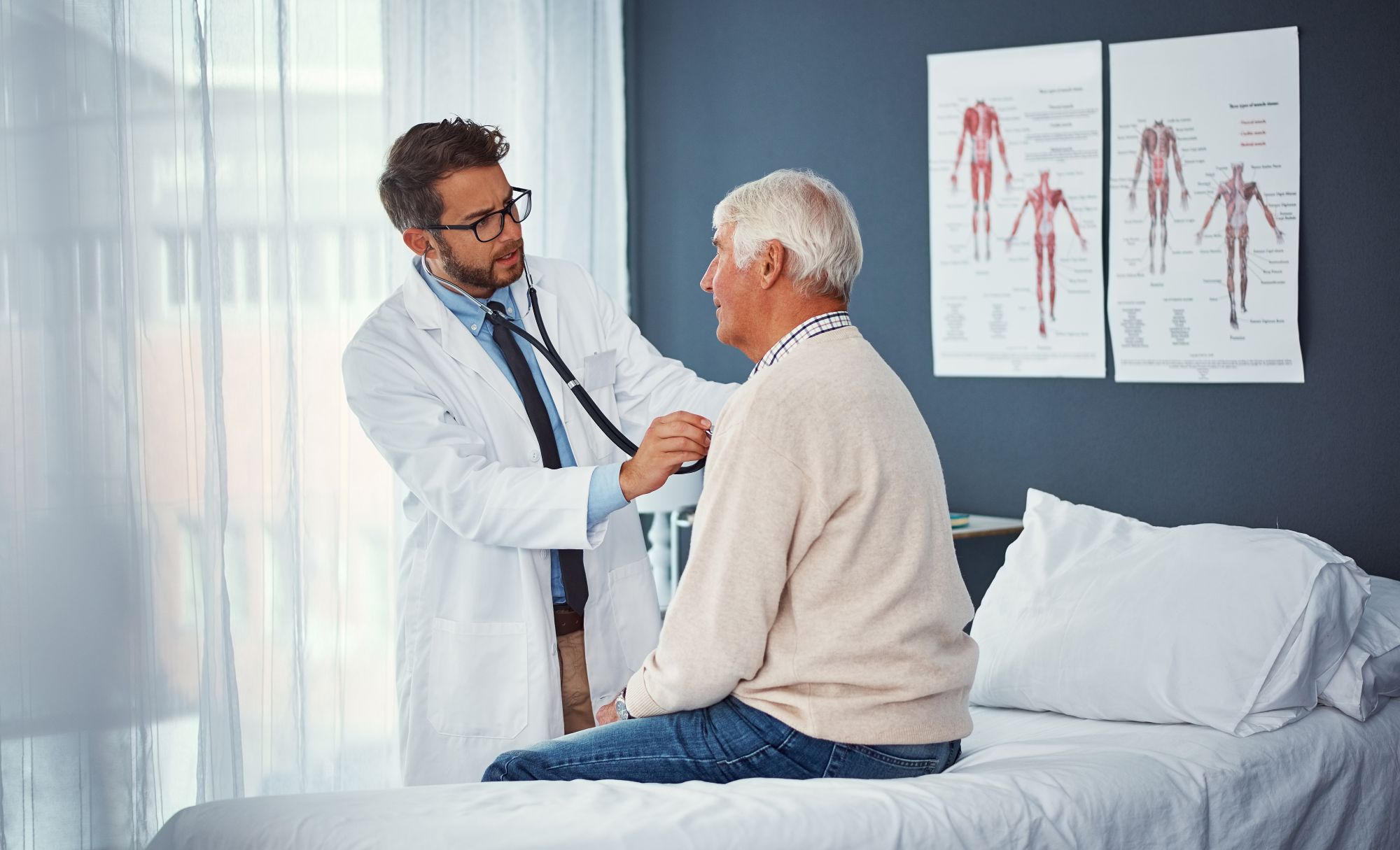 A doctor examines an older male patient in a medical office, with anatomical charts visible on the wall.