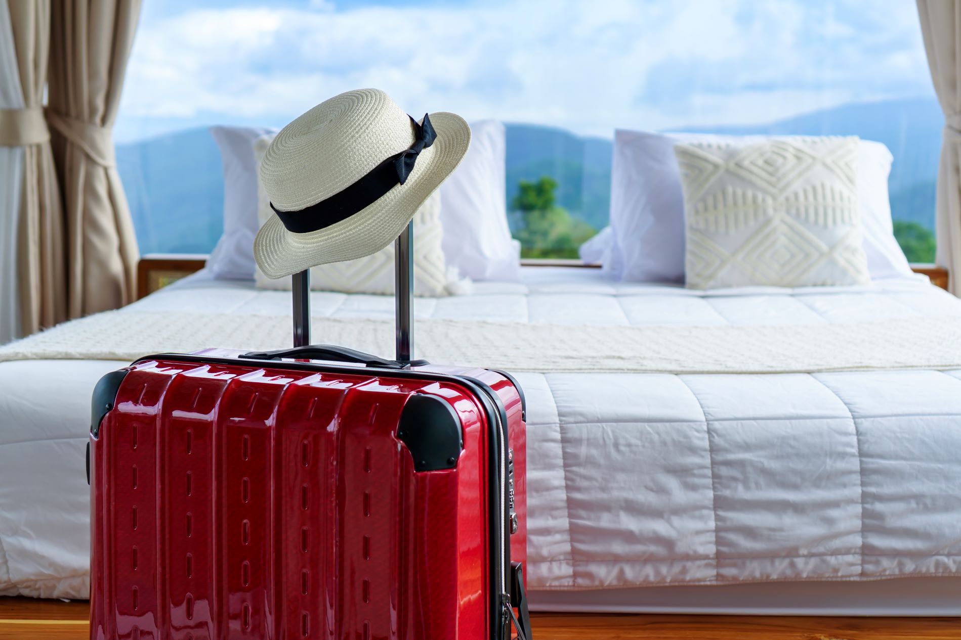 A red suitcase with a straw hat rests beside a bed, suggesting preparations for a memorable trip.