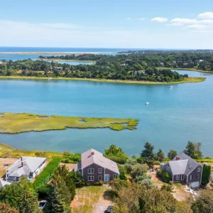 Aerial view of a Cape Cod landscape featuring waterfront homes, lush greenery, and vibrant blue water under a clear sky.